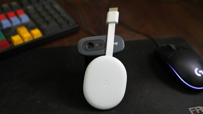 A white Google Chromecast rests atop a web camera, the two items sit between a mouse and keyboard