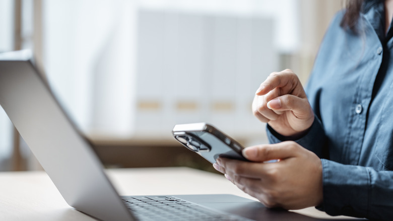 A woman using a smartphone while sitting in front of a laptop computer