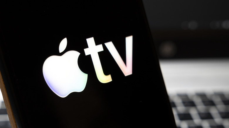 The Apple TV logo is displayed on a smart phone as it sits in front of what appears to be a heavily blurred MacBook