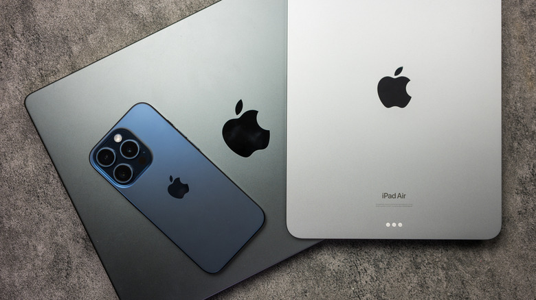 A silver iPad Air and a black iPhone sit on top of a black MacBook laptop, all three devices appears to be on top of a stone background