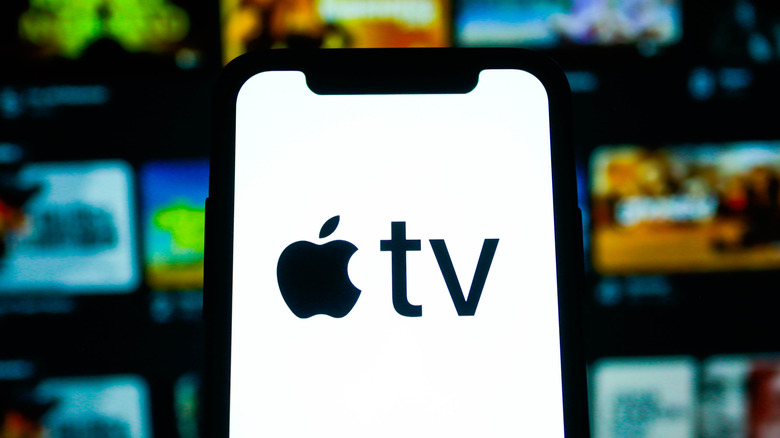 A smartphone displaying the Apple TV logo is in front of what appears to be a heavily blurred shot of the Apple TV Home screen