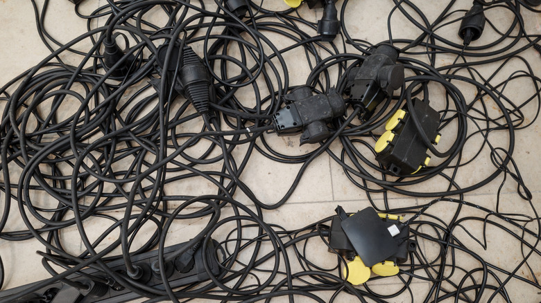 A mess of tangled-up thick black power cords, extension cords, and outlets on a beige tiled floor.