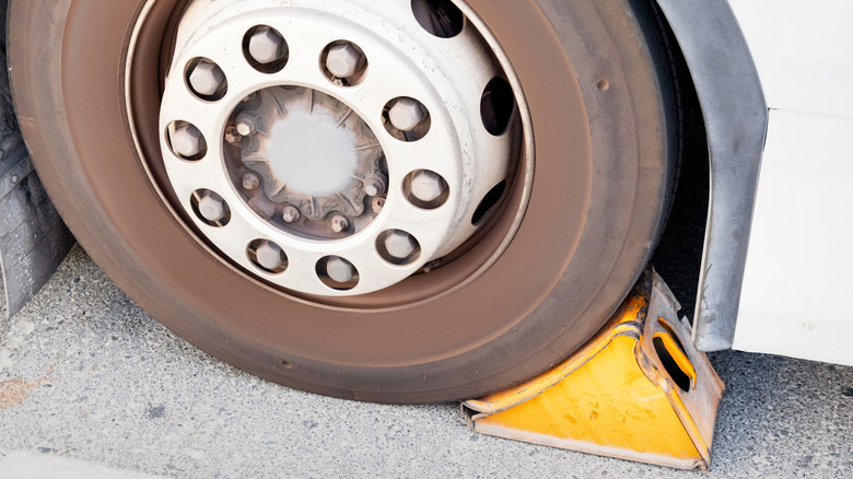 A bright yellow chock behind a large, rusty-looking tire on a gray concrete surface.