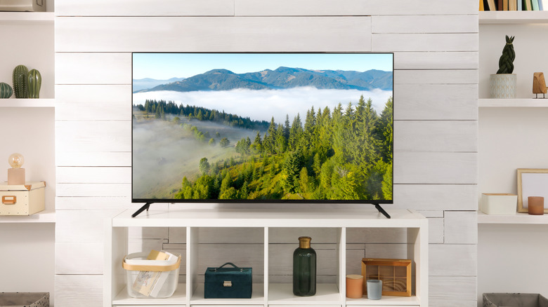 A black television displaying a cloudy mountainside sits atop a white stand in a mostly white room with various objects resting on shelves on each side of the TV