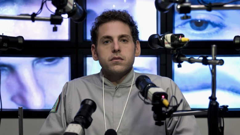 Jonah Hill in the limited 2018 Netflix series "Maniac," Hill is seated at a table surrounded by cameras, behind him are televisions displaying peculiar closeups of his eyes and face