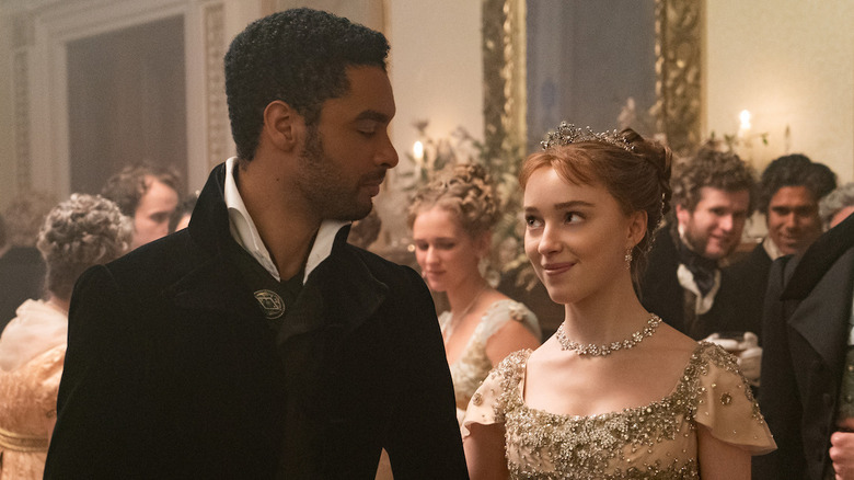 Simon (Regé-Jean Page) and Daphne (Phoebe Dynevor) looking lovingly into one another's eyes while others look on.