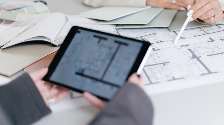 Person using a tablet and stylus while reviewing floor plans and design materials.