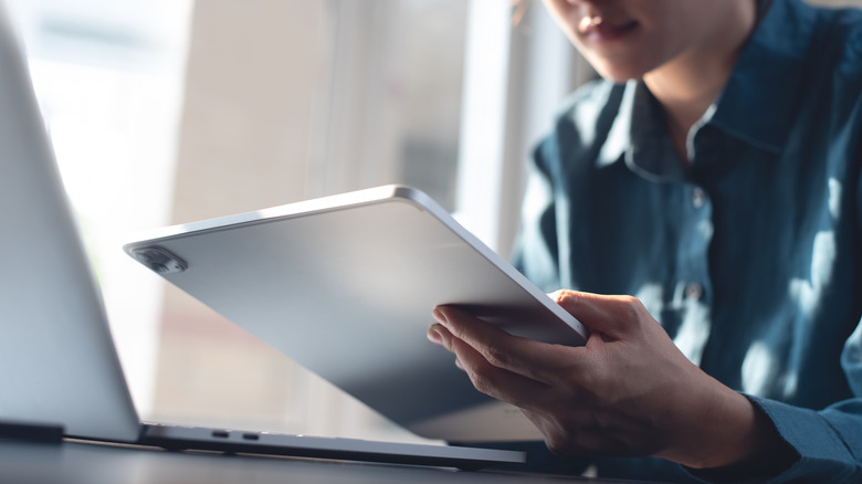 Person holding a tablet beside a laptop on a desk.