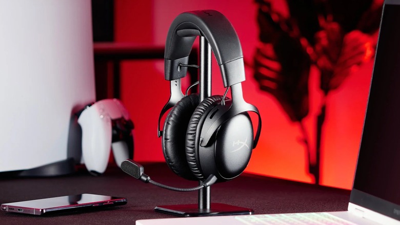 A pair of black HyperX Cloud III S wireless headphones sit on a headphone stand next to a PS5, a smartphone, and a laptop, there is red and orange lighting behind the desk in which all of these object sit