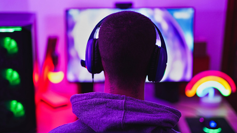 The back of an individual's head as they wear a a pair of gaming headphones and stare at a computer monitor, there are several light scattered throughout the image, as well