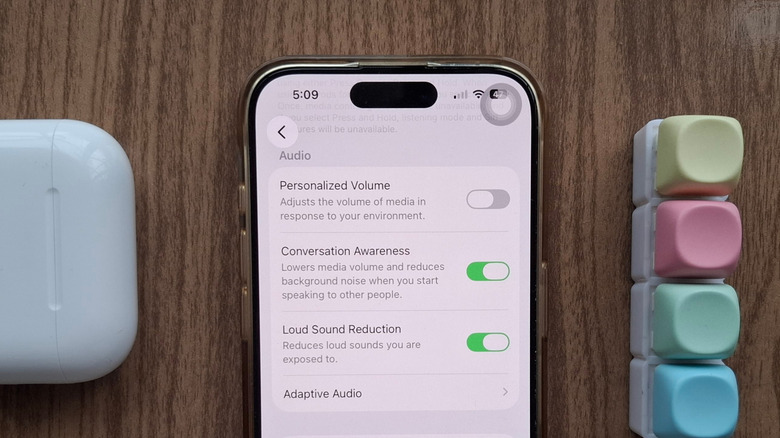 An iPhone showing the Conversation Awareness AirPods setting, next to an AirPods case and a fidget