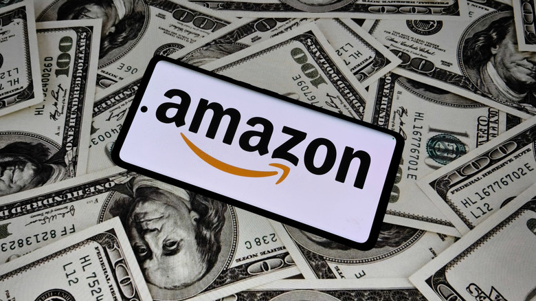 A cellphone displaying the Amazon logo sits atop various hundred dollar bills covering the entire background