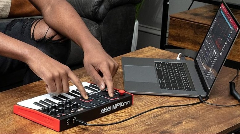 A closeup of a user having both hands on a black Akai MPK Mini MK3 MIDI keyboard controller, the keyboard is connected to a silver laptop by USB
