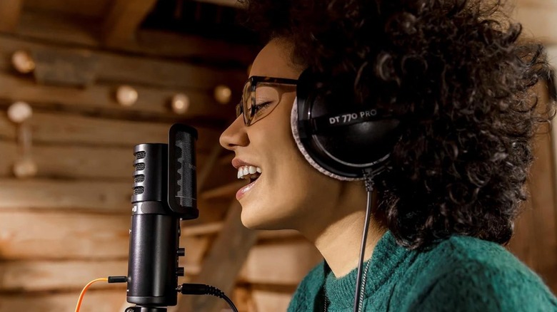 An individual in a green sweater wearing a pair of Beyerdynamic DT 770 Pro Closed-Back Studio Headphones as they sing into a microphone
