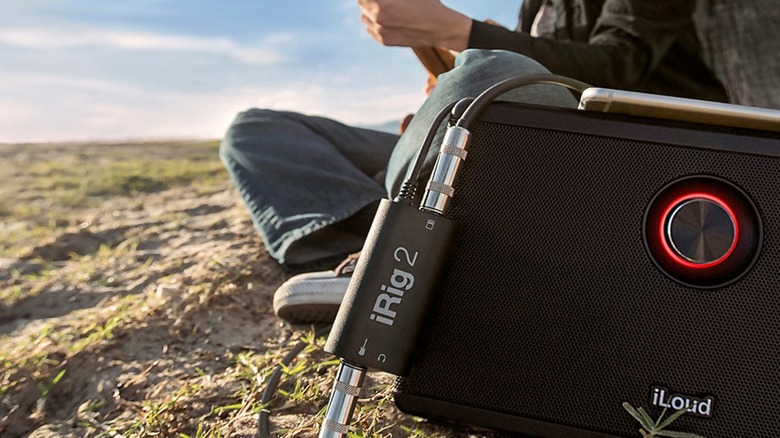 A black IK Multimedia iRig 2 portable electric guitar interface is connected to an iPhone, an outdoor speaker, and an electric guitar that is being held by a player, the photo is shot outdoors