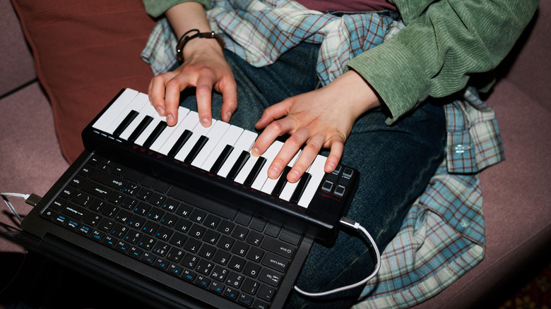 Two hands on a black midi keyboard that's connected via a white USB cable to a black laptop, the person is wearing a blue flannel shirt around their waste and a green shirt