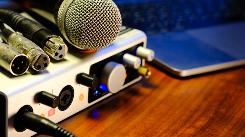 A white audio interface with a microphone and several XLR cables sitting on top of it, the device also sits next to a silver laptop