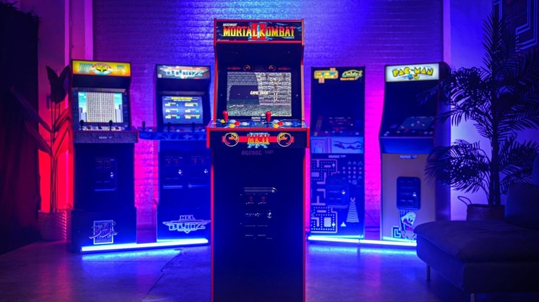 The Arcade1Up "Mortal Kombat II" Deluxe Arcade Machine stands at the center of the image, with four additional arcade machines lit up by lights on the ground standing behind it