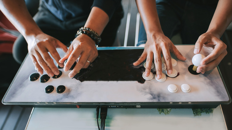 Two individuals sitting behind a white arcade controller, both have their hands on the controller, and the photo features the controller primarily