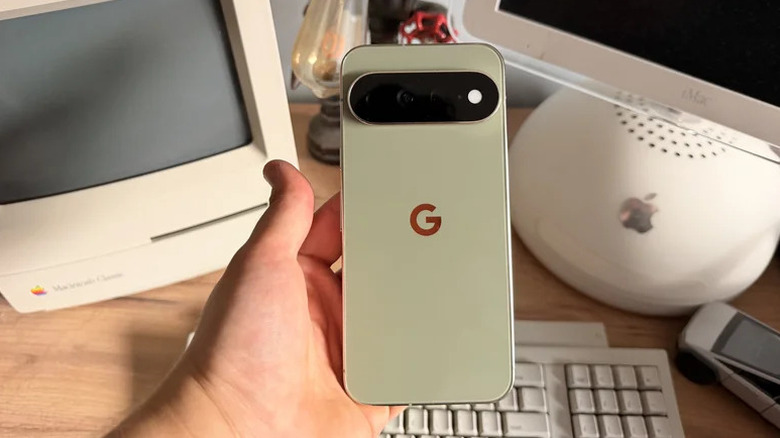 Close-up of a person holding the Pixel 11 Pro XL in Olive color with various gadgets in the background