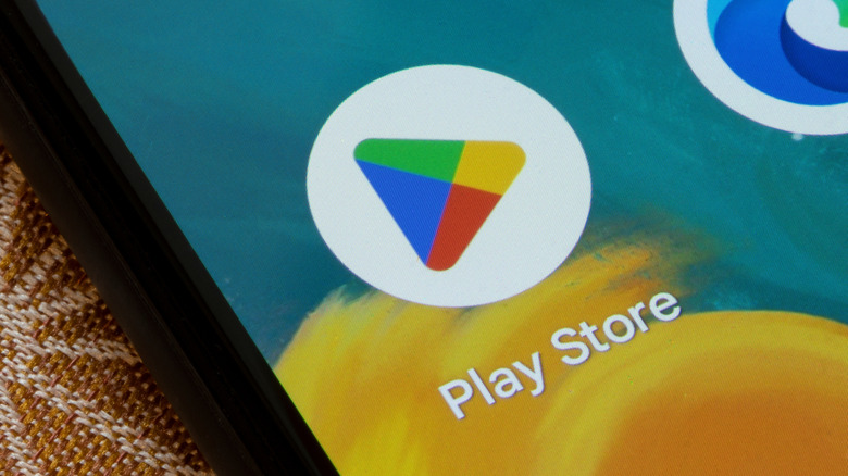 A black smartphone displays the app icon for the Google Play Store, behind the device, it's possibly to see part of the brown patterned fabric the smartphone lays upon