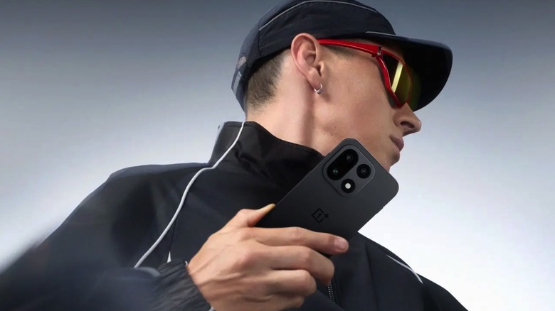 A person wearing a hat and sunglasses with the OnePlus 15 in hand.