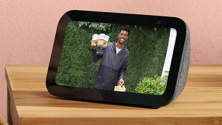 The Amazon Echo Show 5 set up on a bedside table. It shows a live video feed from a camera-equipped doorbell of a man holding coffees and food.