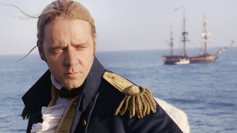 Russell Crowe standing in front of water with a boat behind him in Master and Commander: The Far Side of the World