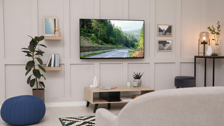 A television is attached to a wall in a primarily white living room, the TV is displaying an open road surrounded by trees