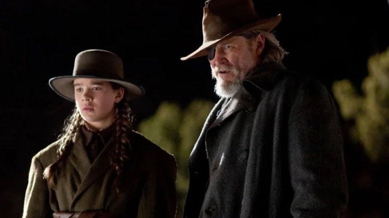 Mattie Ross (Hailee Steinfeld) stood next to Rooster Cogburn (Jeff Bridges) as they both look down at something in True Grit