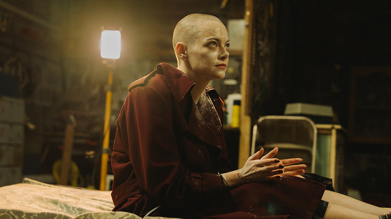 Emma Stone sits upright on a gurney in a dark basement with her head shaved and hands raised in supplication.