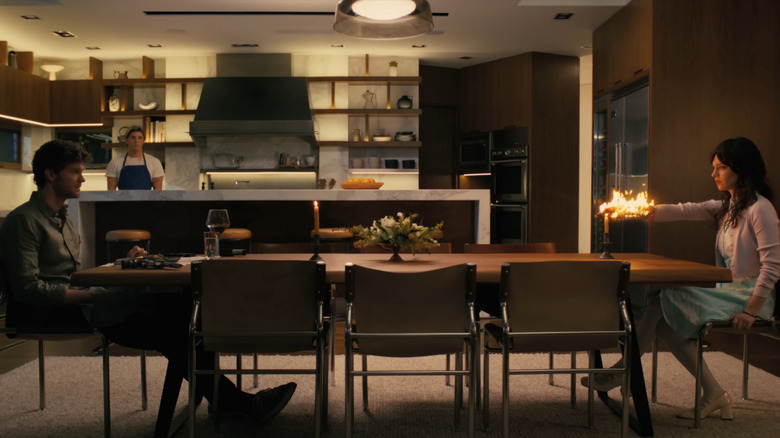 A woman sits across from a man at the dinner table, her hand extended, engulfed in flames, as a chef watches quietly in the background.