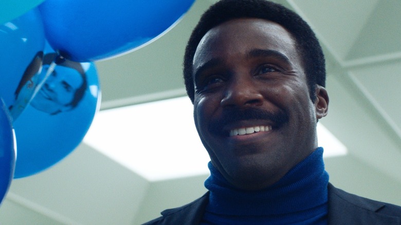 Actor Tramell Tillman in a still from Season 2 of "Severance," the actor is playing Mr. Milchick holding several blue balloons that have the face of Mark S. (Adam Scott) printed on them