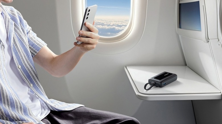 A person using their phone in an airplane with Anker power bank on the tray table
