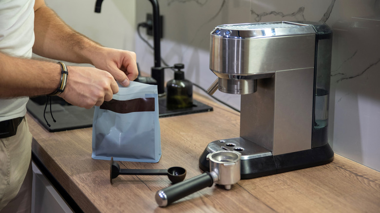 A person opening a bag of coffee grounds next to a coffee machine.