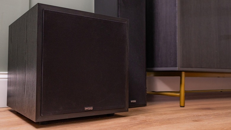 A black Dayton Audio Classic CS1200 subwoofer sits next to another speaker on a wooden floor next to what appears to be part of a table
