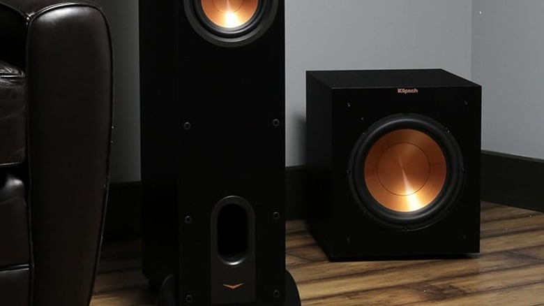 A Klipsch R-12SW subwoofer sits in the corner on a wooden floor, the subwoofer sits next to a tall speaker