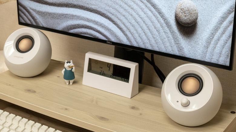 Two white round Creative Pebble speakers on a light wood desk, with a clock and cat figurine between them