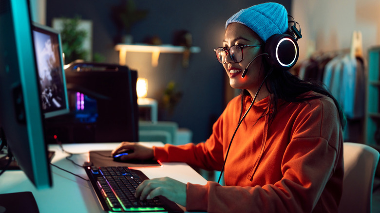 A young woman sitting at a desktop computer playing a video game and wearing headphones