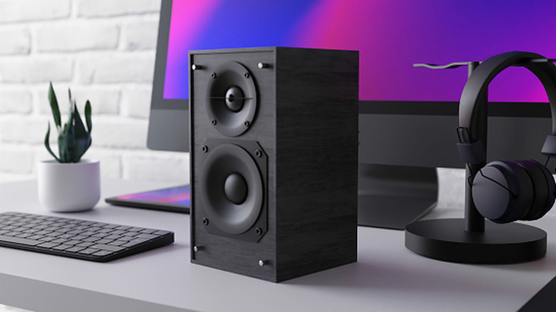 A black speaker on a desk in front of a monitor with headphones and a keyboard on either side