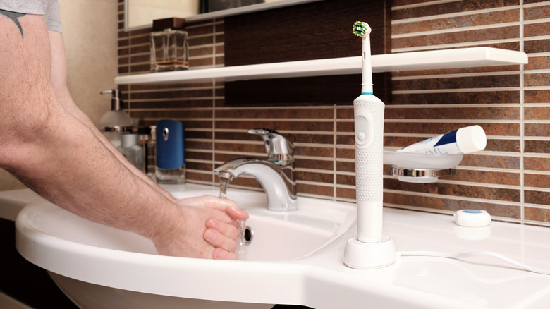 An individual washing both of their hands in the sink, a white electric toothbrush sits vertically next t them on the white bathroom sink