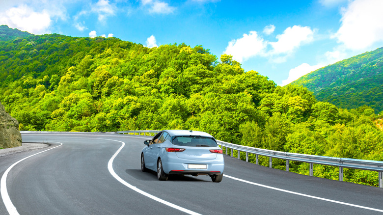 A light blue car driving along a road near a plush forrest landscape