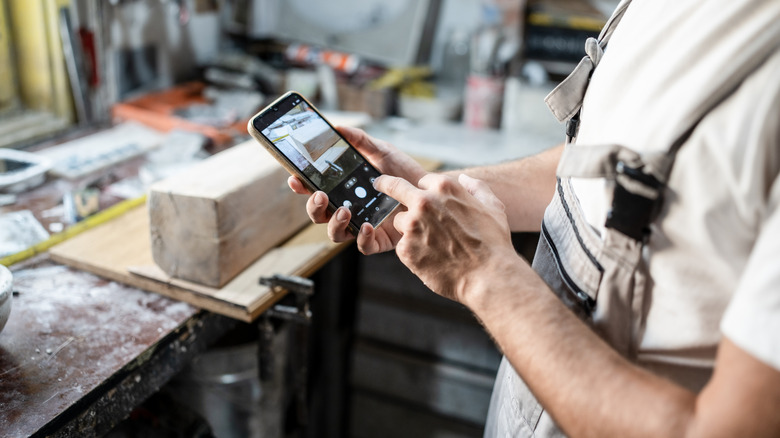 A man in a woodworking shop holding a cell pohone taking a picture of a piece of wood