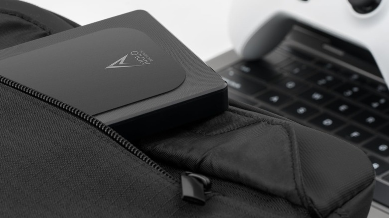 An external hard drive in a bag sticking out the pocket in front of a laptop and video game controller
