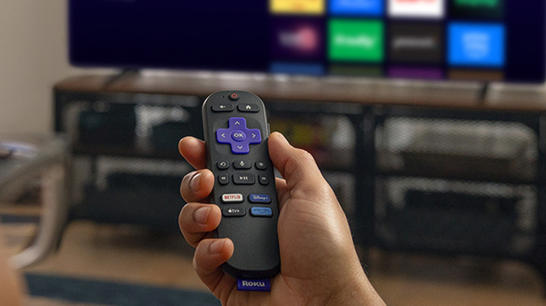 A hand holding the Roku remote with a TV and the Roku menu in the background