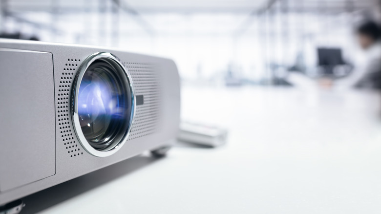 A projector placed on a flat surface with a blurred workplace is the background.