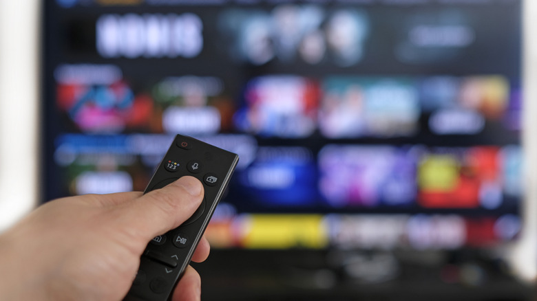 Holding a TV remote with a smart TV seen in the background.