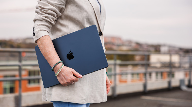 An individual holding a black MacBook in their right arm as they stand outside