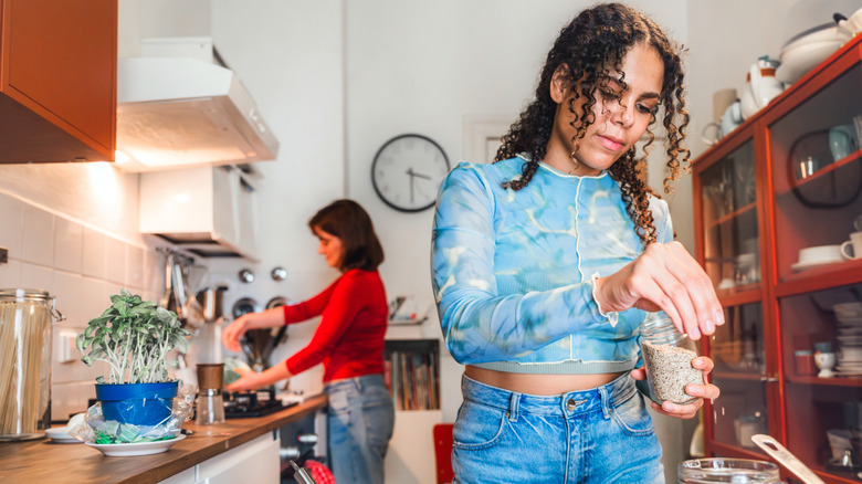 Woman preparing food in a kitchen while another person cooks in the background.