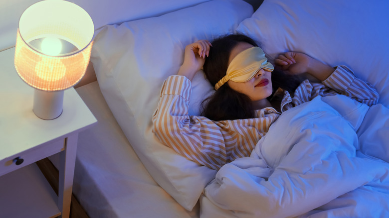 Person sleeping in bed wearing a sleep mask beside a lit bedside lamp.